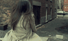 a woman in a trench coat is sitting on the sidewalk in front of a brick building