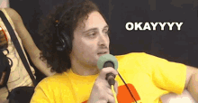 a man wearing headphones and a yellow shirt is holding a microphone and says okayyyy