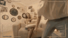 a man is standing in front of a woman sitting on a couch in a living room with clocks on the wall
