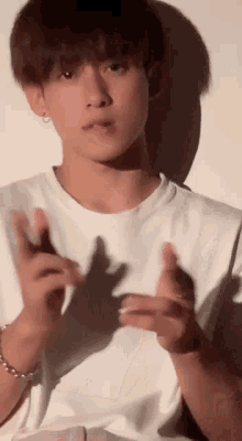 a young man in a white t-shirt is making a hand gesture while standing in front of a white wall .