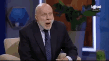 a man in a suit and tie is sitting in a chair with his mouth open while talking on a television show .