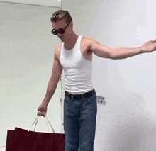 a man in a white tank top and blue jeans is holding a red bag .