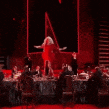 a drag queen in a red dress is dancing on a stage with her arms outstretched .