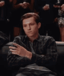 a man in a plaid shirt is sitting on a couch with his arms crossed in front of a crowd of people .