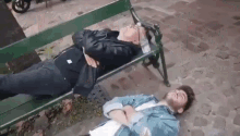 two men are sleeping on a park bench on the sidewalk .