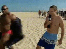 a shirtless man holding a microphone stands on a beach next to another man