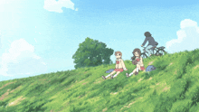 two girls sit on a grassy hill with a man riding a bike