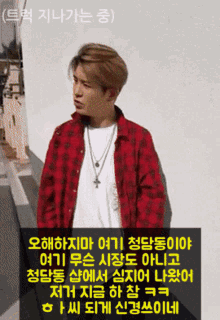 a man wearing a red plaid shirt and a cross necklace stands in front of a white wall