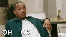 a man in a green jacket is sitting on a couch with his hands on his hips and says `` oh ok '' .