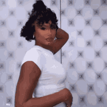 a woman with curly hair is wearing a white shirt and standing in front of a white quilted wall .