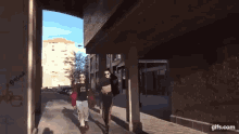 a couple of people walking down a sidewalk with a building in the background and a gifs.com logo