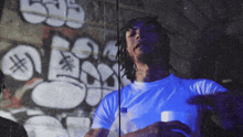 a man in a white shirt is standing in front of a wall with graffiti on it and holding a bottle