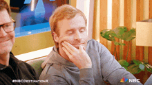 two men are sitting on a couch and one has his hand on his face and the nbc logo is visible in the corner