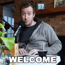 a man sitting at a table with a microphone and a glass of wine with welcome written on the table