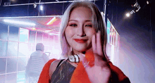 a woman with blonde hair is waving at the camera in a dark room while wearing an orange jacket .