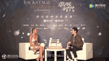 a woman sits in a chair talking to a man in front of a backstage secrets sign