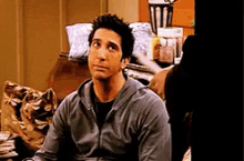 a man in a grey sweatshirt is sitting on a couch looking at the camera .