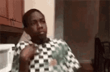 a man in a checkered shirt is sitting in a kitchen with his hands in his pockets .