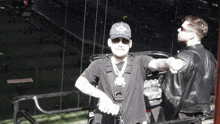 a man wearing sunglasses and a hat with the letter o on it stands next to another man in a leather jacket
