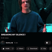 a man in a blue sweater is standing in front of a tree and says breaking my silence
