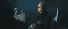 a bald man in a black jacket stands in a dark room with a statue in the background