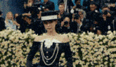 a woman is walking down a runway in front of a crowd of people wearing a hat and pearls .