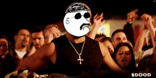 a man with a cross necklace stands in front of a crowd with $ dood written on the bottom right