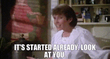 a woman is sitting in a kitchen with the words `` it 's started already look at you '' written on the screen .
