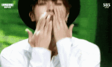 a man wearing a hat and a white shirt is covering his mouth with his hands .