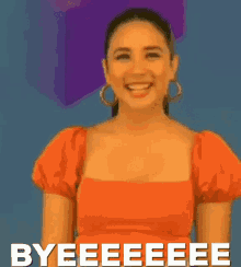 a woman in an orange top is smiling and says byeeeeee