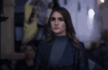 a woman wearing a black jacket and a grey turtleneck looks at the camera