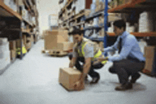 a man is squatting down to lift a box in a warehouse while another man looks on .