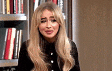 a woman with long blonde hair is sitting in front of a bookshelf in a room .