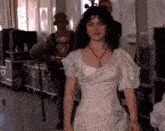 a woman in a white dress with a red necklace stands in front of a camera