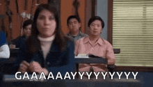 a woman is sitting at a desk in a classroom with other people and making a funny face .