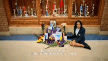 a girl sits on the floor in front of a display of trophies and flowers and says " girl we 're with you "