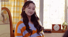 a woman in a striped sweater is smiling in a room with a clock on the window .