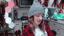 a woman wearing a beanie and a plaid shirt is in a kitchen