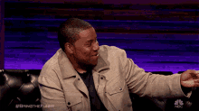 a man in a tan jacket is sitting on a couch with a nbc logo in the background