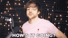 a man in a pink shirt stands in front of a microphone with the words how 's it going