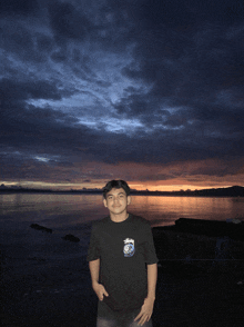 a man wearing a stussy shirt stands in front of a sunset over a body of water