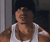 a man wearing a beanie , a tank top , and a gold chain is looking at the camera .