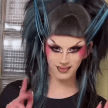 a drag queen with long black hair and blue highlights is making a peace sign .