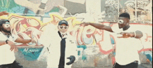 a group of men wearing masks are dancing in front of a wall with graffiti on it