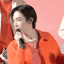 a man in an orange shirt is singing into a microphone while wearing a teddy bear necklace