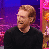 a man with a beard wearing a black shirt smiles in front of flowers