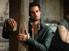 a man in a green jacket leans against a wooden pillar