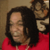 a man with dreadlocks and a beard is wearing a red shirt and looking at the camera .