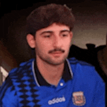 a man with a mustache wearing a blue adidas shirt is sitting in a chair .
