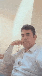 a man wearing glasses and a white shirt is sitting down with his hand on his chin .